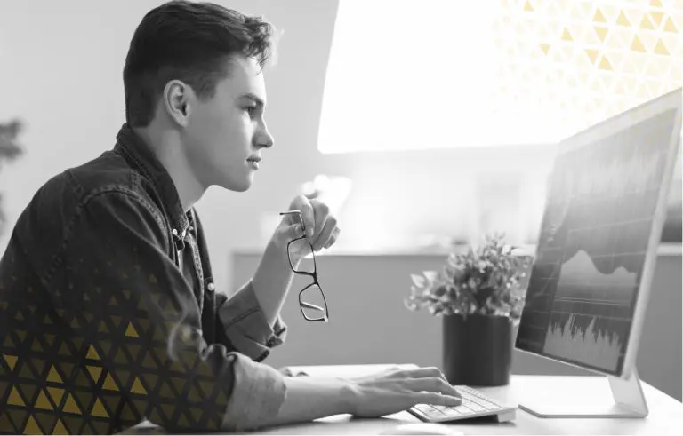 guy sitting at desk holding glasses looking at a computer